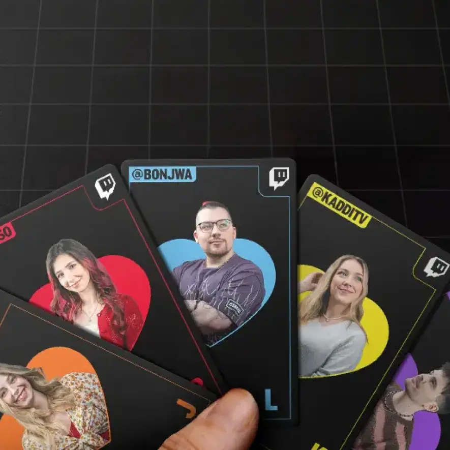 Eine Hand hält aufgefächerte, schwarze Spielkarten. Auf den Karten befinden sich Porträts von Twitch-Streamern in bunten Herzen sowie deren Benutzernamen, darunter @BONJWA und @KADDITV, zusammen mit dem Twitch-Logo.